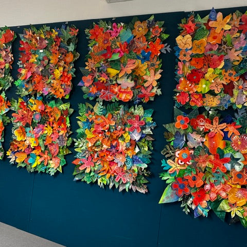 Eight canvases covered in layered paper flowers and leaves made by students at Omokoroa No 1 School, mounted together on a teal wall