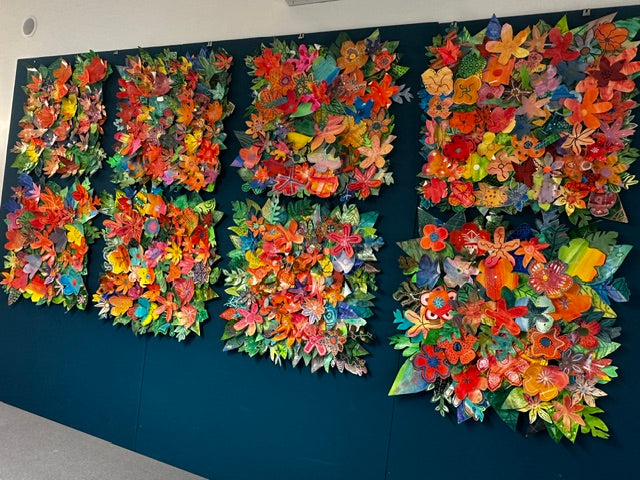 Eight canvases covered in layered paper flowers and leaves made by students at Omokoroa No 1 School, mounted together on a teal wall